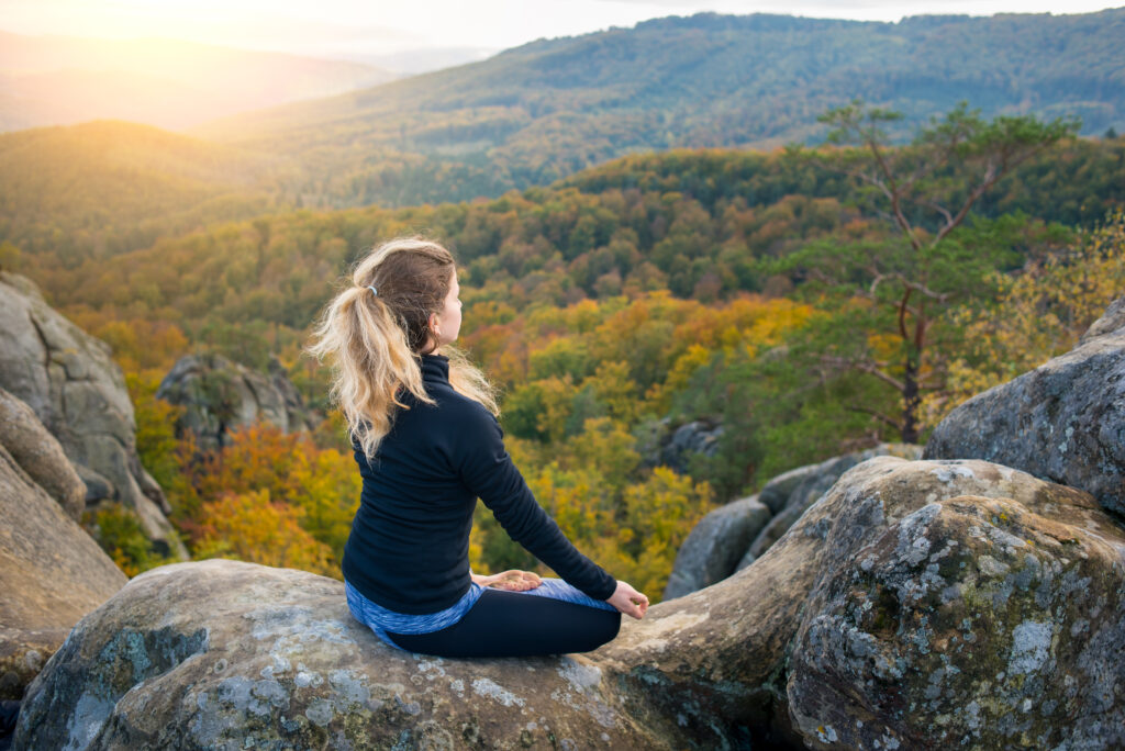 Peaceful fit female is practicing yoga and doing asana Siddhasana on the top of the high rocky mountain in the evening. Beautiful sunset, autumn forests, rocks and hills on the background