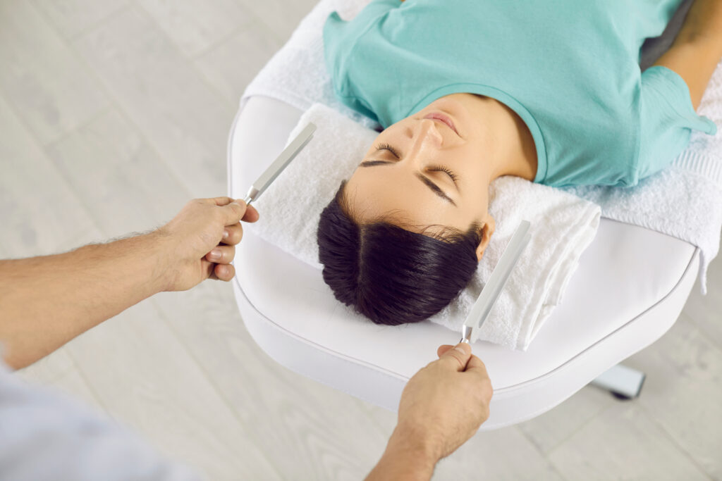 Sound therapy specialist working with patient. Healer holding two metal tuning forks near young lady's head. Holistic music healing, relaxation, stress reduction, restoring inner balance concept