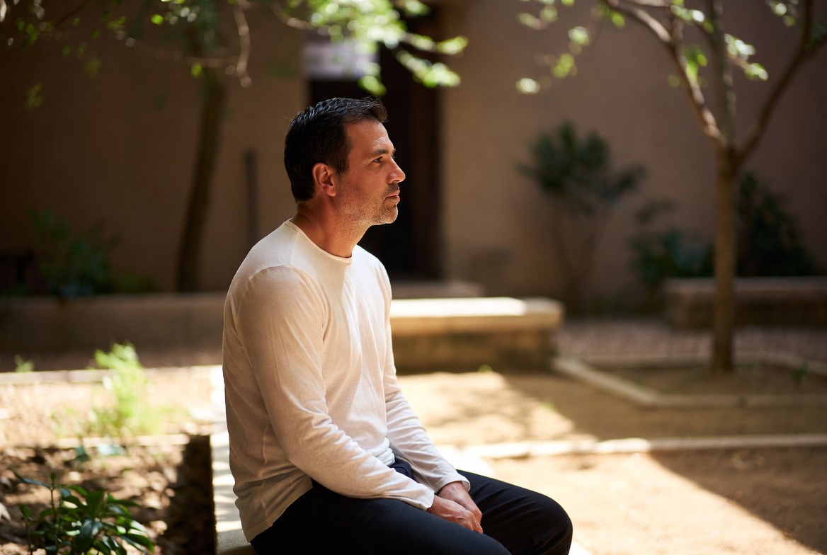 A man sitting alone in a sunlit courtyard, looking off to one side